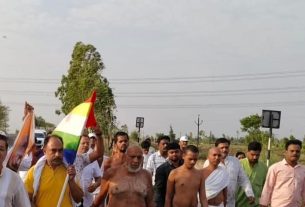 Group of men marching on a rural road, one person holds a rainbow flag high overhead.