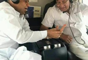 Two men in headsets sit in a small aircraft cabin, one in white traditional clothing showing something on a small orange object to the other as they talk.