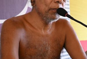 A shirtless older man with a gray beard speaks into a microphone at a public event, seated at a table with a colorful backdrop behind him.