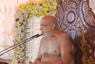 A bald, bearded elderly man sits shirtless on a large ornate wooden throne, speaking into a microphone at a flower-decorated podium.
