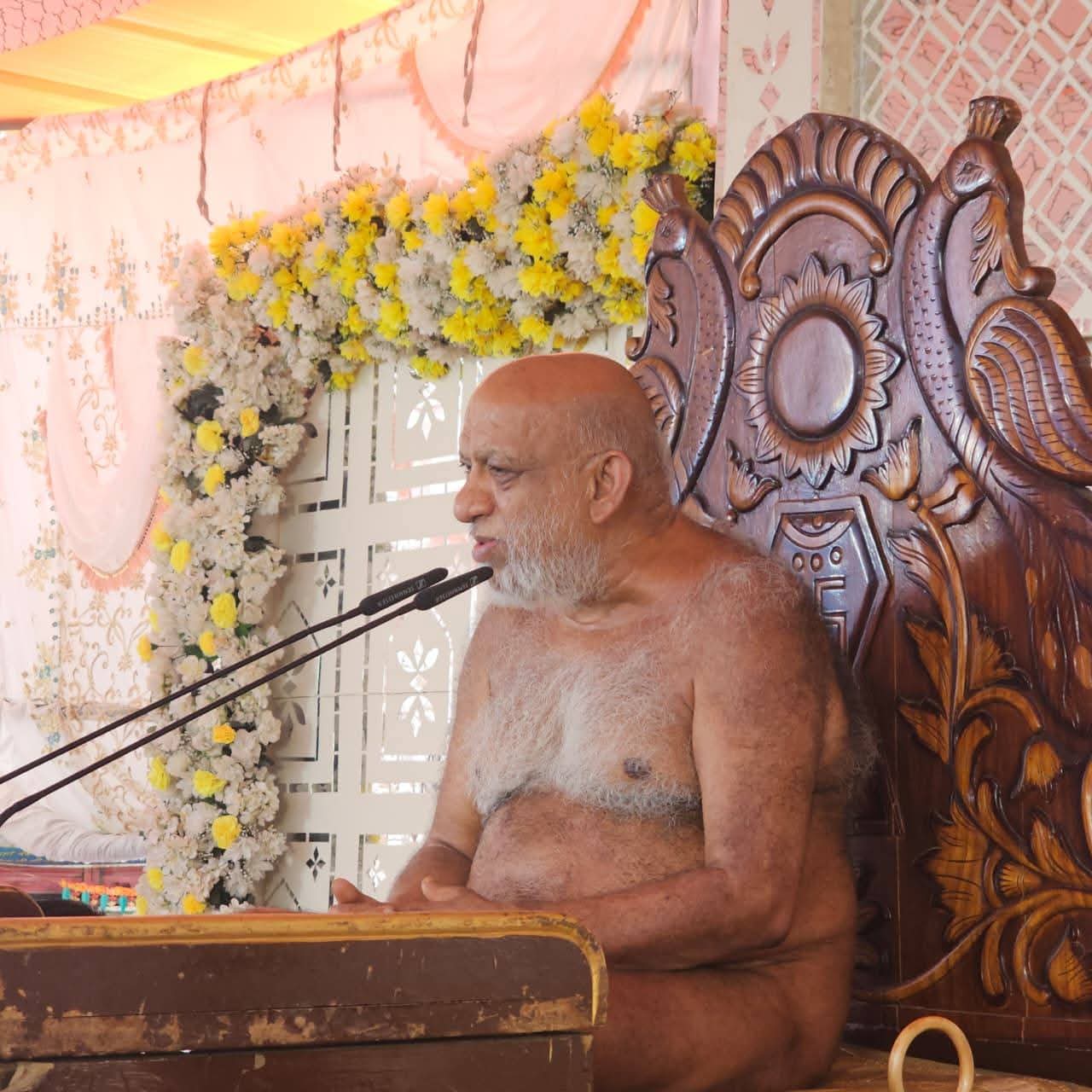 A bald, bearded elderly man sits shirtless on a large ornate wooden throne, speaking into a microphone at a flower-decorated podium.