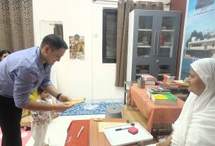 Man bending to hand a book to a little girl in a room filled with books and furniture; woman in white hijab sits nearby smiling.