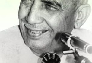 Smiling man in a white cap speaks into a microphone during a public address with a vintage setup.