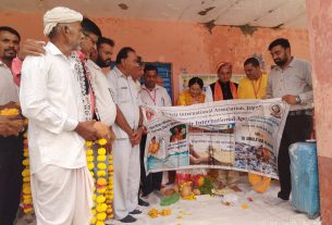 Group of Indian men on a stage holding a large banner at a community event, wearing garlands of marigolds against a pink wall backdrop.