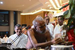 Bearded elderly man performing a temple ritual, presenting a small silver statue to a stand while several men in white robes observe in a lit ceremonial hall.