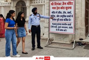 Police officer points to a large rules sign outside a white marble temple with intricate carvings. The sign lists visitor rules in Hindi and dress-code details while onlookers stand nearby.