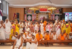 Large group of Hindu devotees in white and saffron robes posing and clapping at a ceremonial hall with an ornate stage backdrop of red, gold, and green decorations.