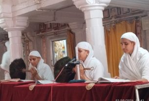 Men in white robes with white head coverings sit at a red-draped table with microphones at a temple ceremony.