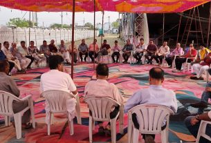 Group of people seated in a circle under a colorful canopy at an outdoor community meeting.