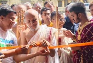 Group of people at a ribbon-cutting ceremony, a man in white traditional robes ties the orange ribbon as onlookers watch.