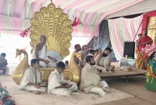 Monks and attendees sit on a decorated stage under pink drapes, with a large golden peacock throne and colorful flowers nearby.