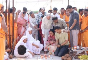 Group of people gathered for a Hindu ceremonial ritual outdoors; priests in white lamps light clay lamps while devotees pray nearby, with orange-robed monks observing.