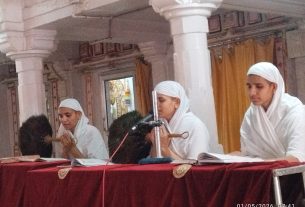 Three people in white robes and turbans sit at a red-draped table inside a temple, with one speaking into a microphone.