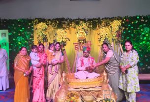 Group of people standing on a decorated stage around a seated man in ceremonial attire with a flower-draped backdrop.