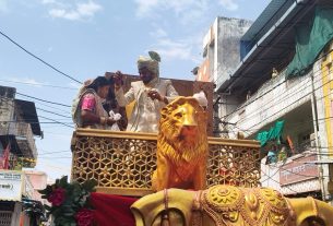 Bride and groom in traditional Indian wedding attire on a decorated float with a gold lion statue in the foreground.