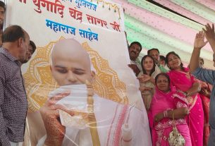 Group of men and women celebrate beside a large banner featuring a bald man holding a ceremonial staff, under a decorated tent backdrop including pink and green fabric patterns.