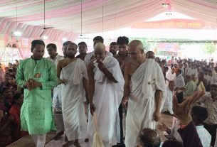 Men in white traditional robes walk through a crowd at a festival under a pink striped canopy; a man in green kurta watches on the left and others smile. This appears to be a ceremonial procession with spectators surrounding the path.