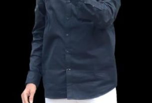 A man with glasses and a gray beard wearing a navy button-down shirt and white pants, speaking into a handheld microphone against a black background.