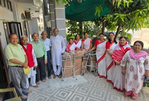 Group of elderly men and women posing outside a building, with walkers and boxed donated goods nearby under a green canopy.