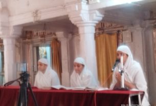 Three men in white robes and head coverings sit at a table with microphones in a marble columned room, likely performing music or chants.