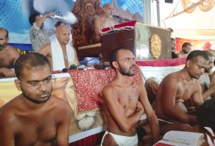 Group of shirtless men wearing white loincloths seated on a decorated stage, with a wooden podium and a blue banner in the background.