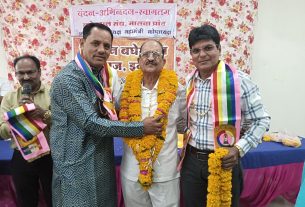 Three men pose at a celebration; center man in white is garlanded with marigolds, flanked by two men wearing rainbow sashes, floral backdrop behind them.