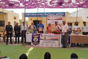 Group of people standing behind a table with posters and banners at an outdoor event under a purple decorative canopy. (India)