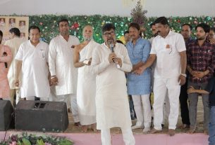 Man in a white kurta speaks into a microphone on a decorated stage, surrounded by men in traditional attire.