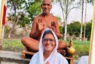 Smiling woman in a white headscarf and robe sits with hands in prayer in front of a shiva-like guru figure meditating outdoors.