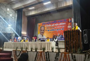 Stage event with a large orange banner featuring a portrait and Hindi text; a man at a podium wearing a yellow shawl speaks, flanked by a panel seated at a long table adorned with flowers and garlands, audience in front.