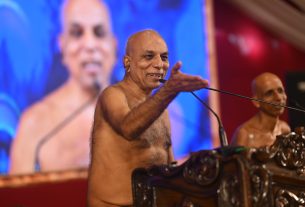 Shirtless man at a carved wooden podium speaking into a microphone and gesturing with his hand on a blue-lit stage
