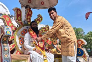 Two men in traditional attire on a bright, ornate festival float; one man places a flower garland on the seated participant.