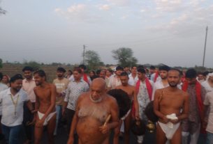 A large group of men walking together in a rural road procession, led by an elderly bearded man holding a wooden staff.