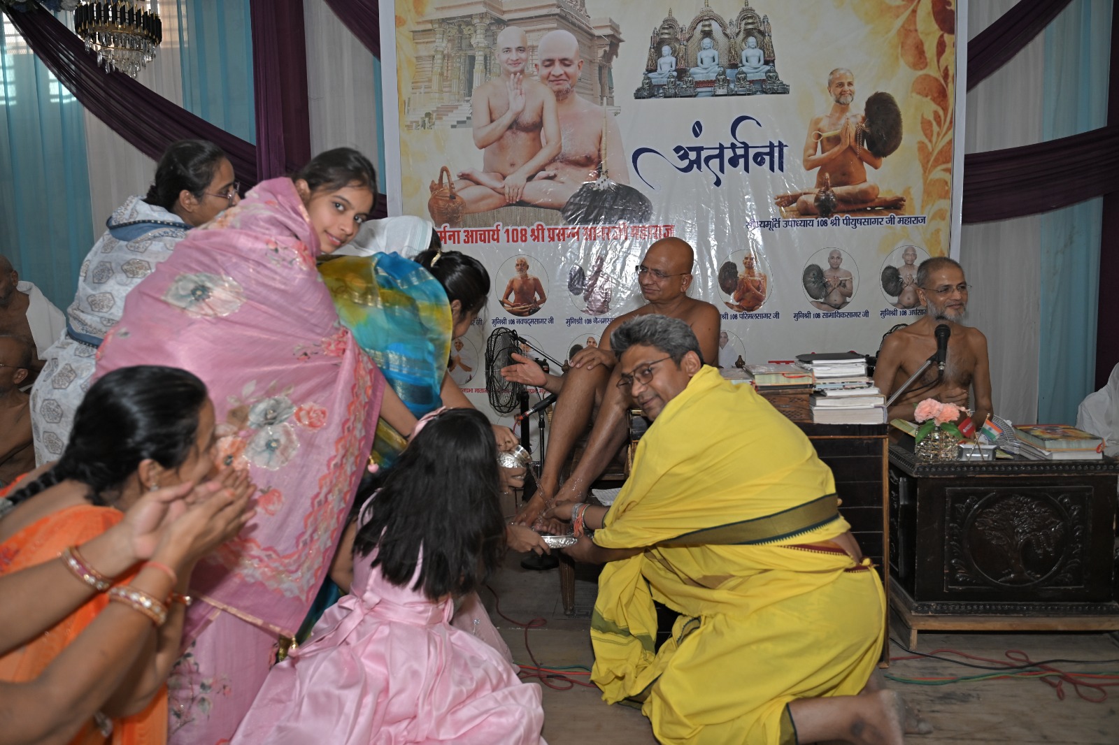 Monk in yellow robes blessing kneeling participants during a ceremony in front of a banner with Hindu imagery.