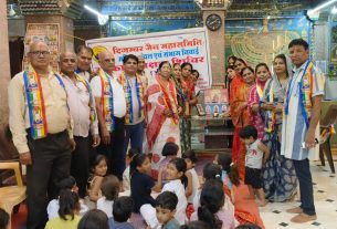 Group of adults wearing colorful sashes posing for a photo inside a temple hall, with children seated on the floor in front of them and a banner in the background.