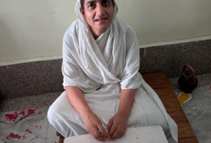 Woman in white traditional robes sits cross‑legged on a low bench, hands folded, with a peacock feather display on a white surface in front and 'Om Shanti' written on the wall behind.