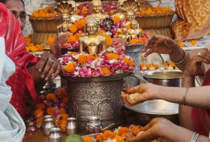 Devotees offering flowers at a religious altar: a large seated golden Buddha statue with a halo, surrounded by smaller statues, rangoli of petals, and metal vessels; people in saffron and colorful attire place flowers and pour offerings as part of a ritual.