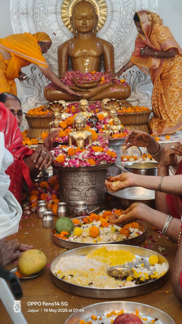 Devotees offering flowers at a religious altar: a large seated golden Buddha statue with a halo, surrounded by smaller statues, rangoli of petals, and metal vessels; people in saffron and colorful attire place flowers and pour offerings as part of a ritual.