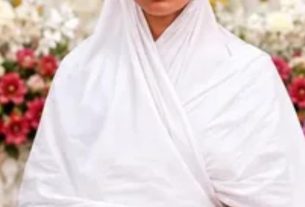 Woman in a white robe and headscarf meditating with eyes closed in a flower-adorned interior, hands resting softly in lap.