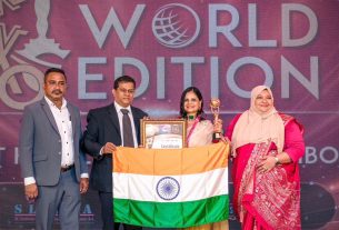 Award ceremony on stage with four people posing; a woman in beige sari holds a framed certificate and trophy, standing behind an Indian flag. backdrop reads '4th Edition Asia Cake Oscars World Edition'.
