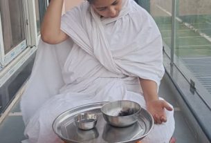 Person in white robes sits cross-legged on a balcony, hand on head, with a metal tray and two bowls on a small yellow stand in front.