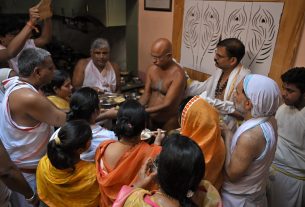 Devotees gather around a shirtless priest performing a Hindu ritual with metal bowls of offerings in a temple kitchen area.