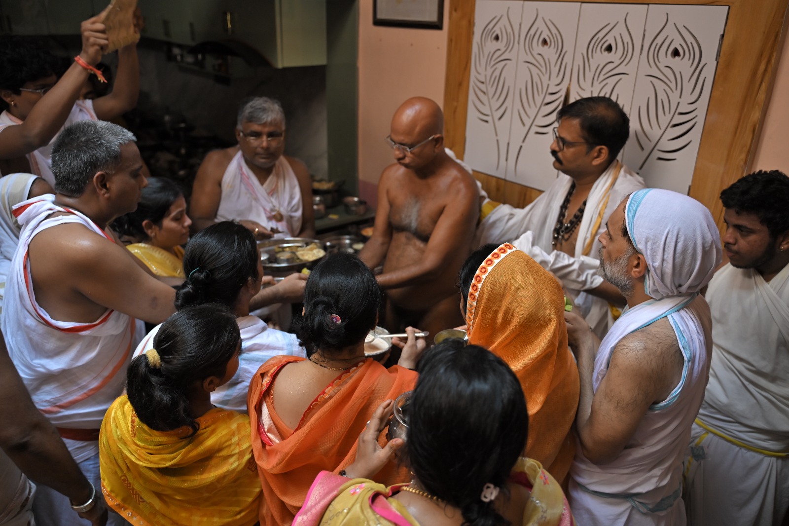 Devotees gather around a shirtless priest performing a Hindu ritual with metal bowls of offerings in a temple kitchen area.