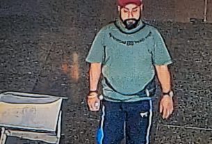 Bearded man in a teal shirt and red cap walks barefoot on a dark street beside a box labeled Fragile.