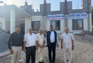 Five men in shirts and jackets pose in front of a partly built brick building with banners in Hindi above the windows and a gravel yard behind them.
