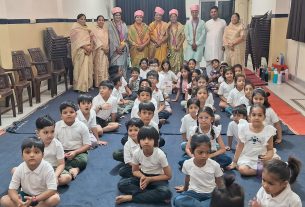 Group of smiling young students sitting cross‑legged on a blue carpet in a school hall, with teachers in traditional attire standing at the back for a ceremony.
