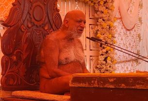 Elderly man with a white beard sits shirtless on a carved wooden throne at a floral stage, speaking into multiple microphones.