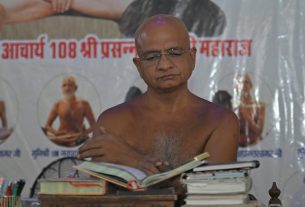 Monk with glasses reading a book at a cluttered desk, purple powder on shoulders, backdrop of a banner with Hindi text behind him