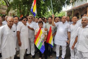 Group of mostly men in white outfits posing with flags at a daytime outdoor gathering; a young boy stands at center holding flags similar to the national colors.