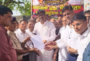 Group of men, mostly in white shirts, exchange a document at an outdoor event; a police officer stands nearby as a colorful banner in Hindi looms behind.